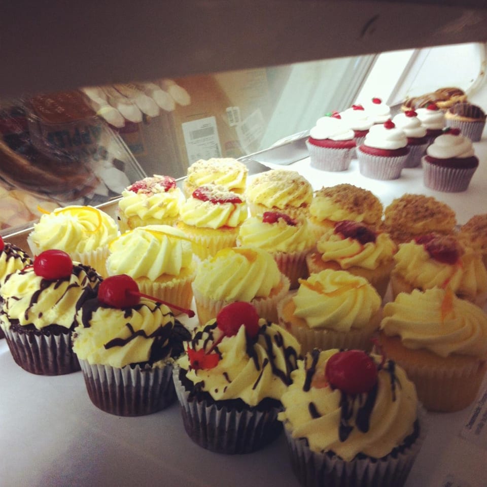 A display case filled with rows of various cupcakes, including red velvet and chocolate with cherries.