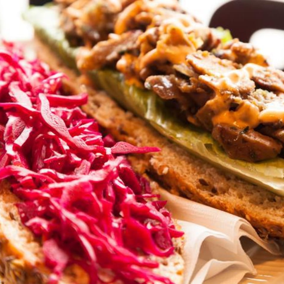 Close-up of a Brooklyn Standard sandwich with meat, sauce, pickles, and red cabbage on bread.