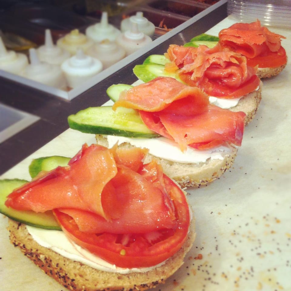 Four bagels with cream cheese, smoked salmon, tomato, and cucumber slices on a counter.