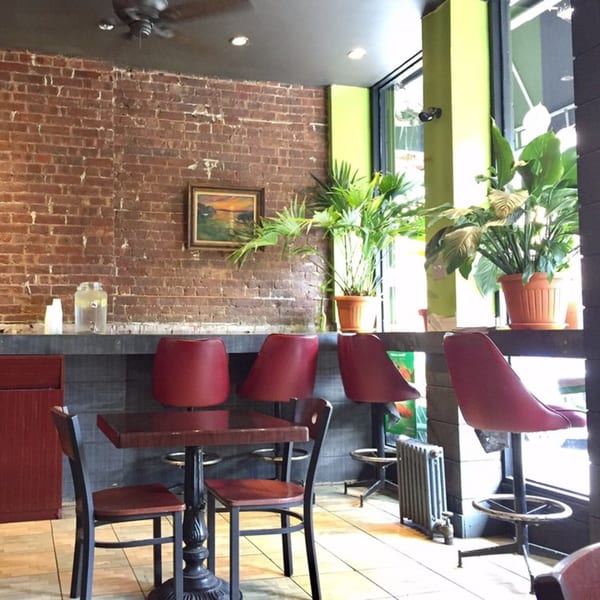 Interior of Brooklyn Standard with brick walls, red bar stools, a small table, and potted plants.
