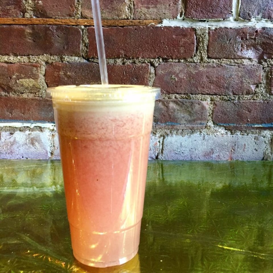 A pink smoothie with a straw in a plastic cup sits on a green table against a brick wall.