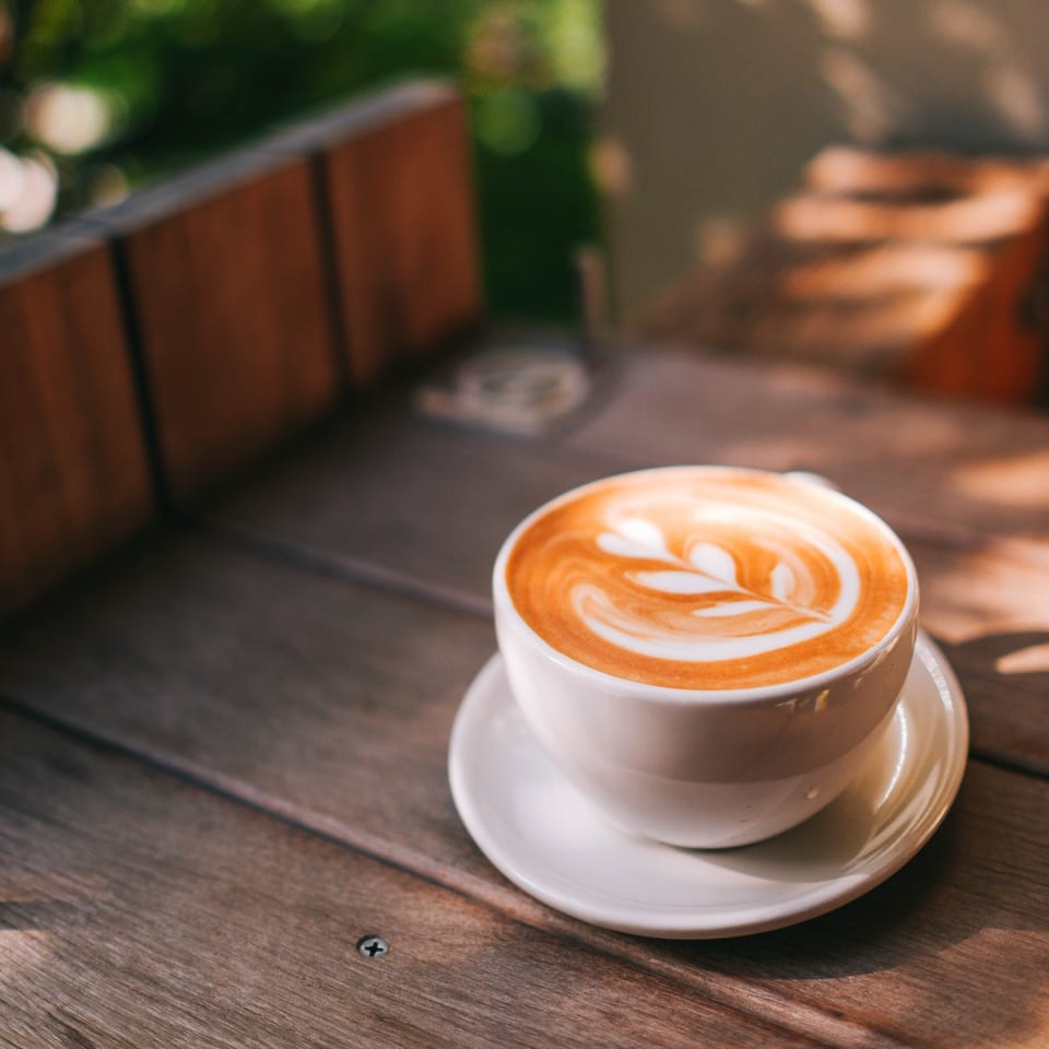 A white cup of latte with leaf art sits on a wooden table in sunlight.